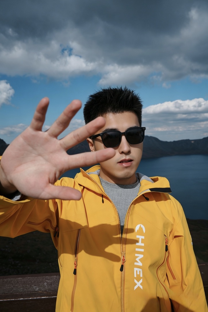 Joey 在旅行途中，身穿黄色外套站在山湖前